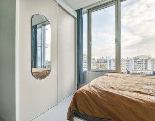 A bedroom in a high rise apartment, a mirror on the wall reflecting the view from the large window next to the bed.