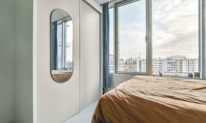 A bedroom in a high rise apartment, a mirror on the wall reflecting the view from the large window next to the bed.