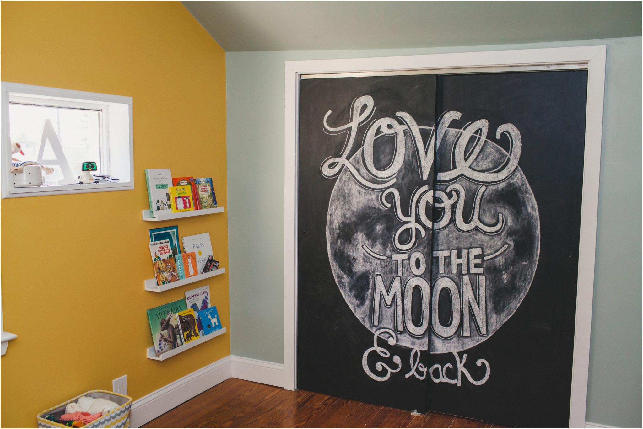 Space children's room with a yellow wall with hanging shelves and a chalkboard that reads "Love you to the moon & back."