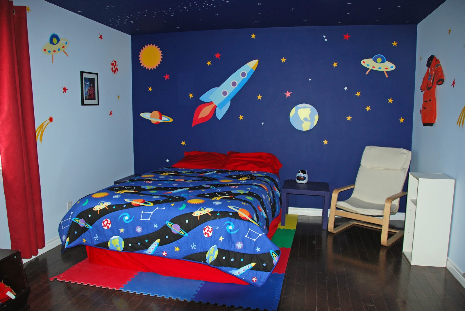 Bed on top of foam squares flanked by space wall art the includes a rocket, UFO, Earth, and the sun.
