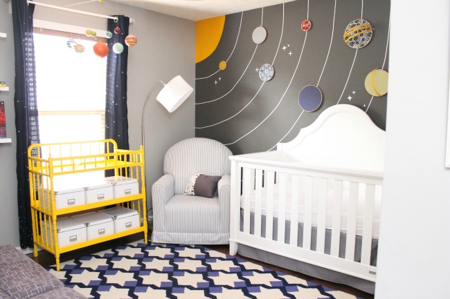 Space nursery with solar system wallpaper, white crib, and a yellow changing table.
